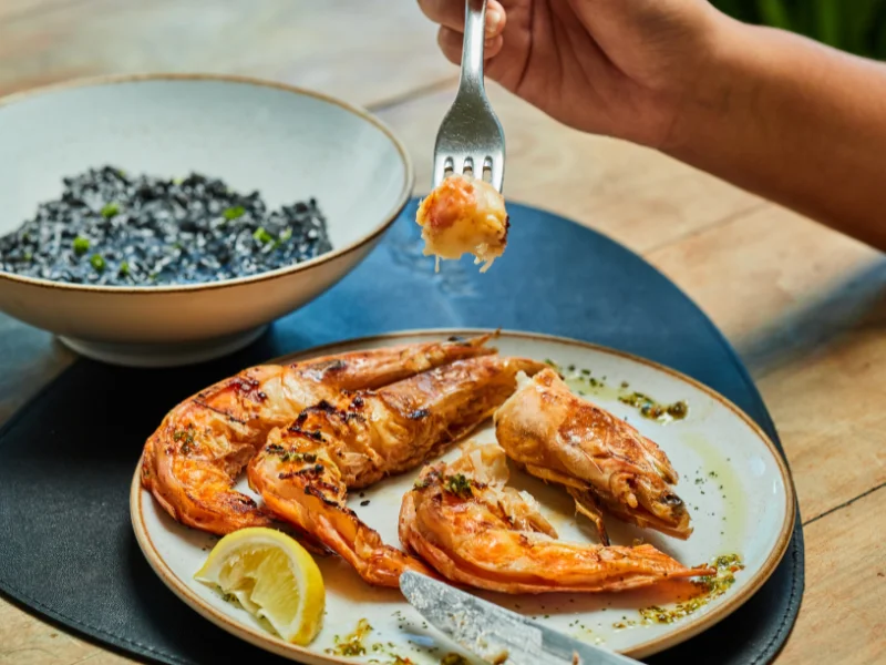 Camarões grelhados inteiros servidos com limão e uma tigela de risoto de arroz negro ao fundo.