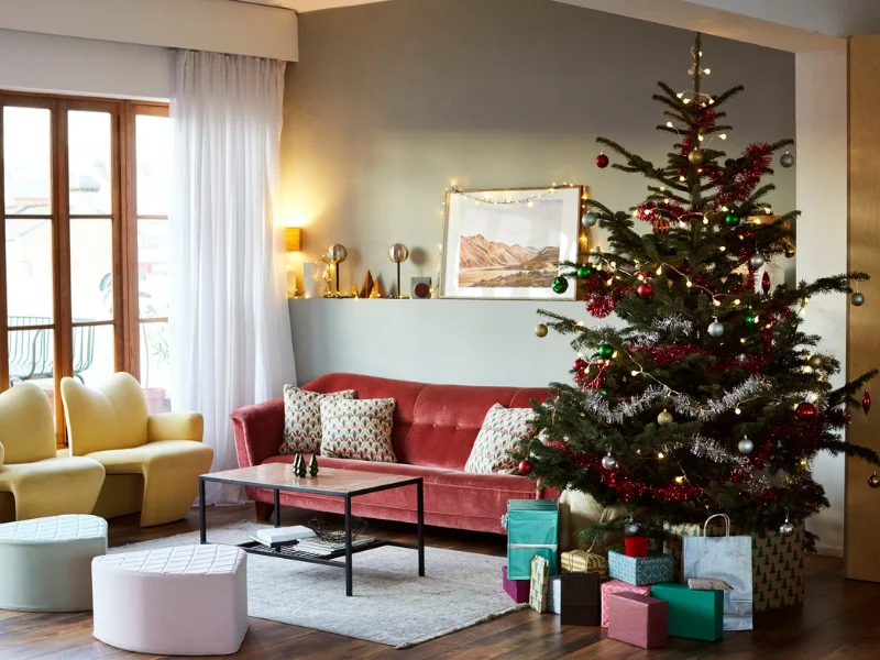 Vista de uma sala de estar moderna e aconchegante com uma grande árvore de Natal decorada e cheia de presentes embrulhados na base. A sala tem um sofá de veludo rosa, duas poltronas amarelas, e luz natural entrando por uma janela à esquerda.