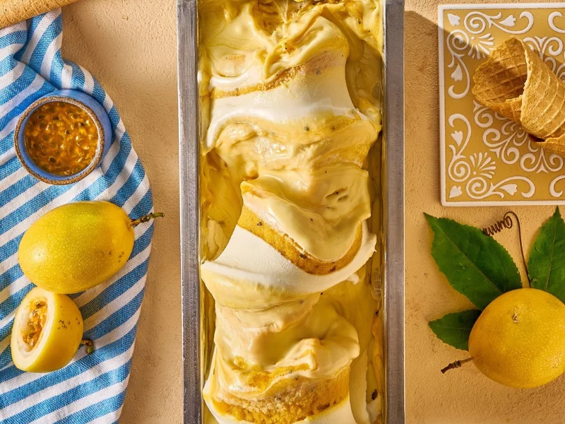 Gelato de maracujá em uma cuba metálica com mesclas da fruta, cercado por maracujás frescos e casquinhas crocantes.