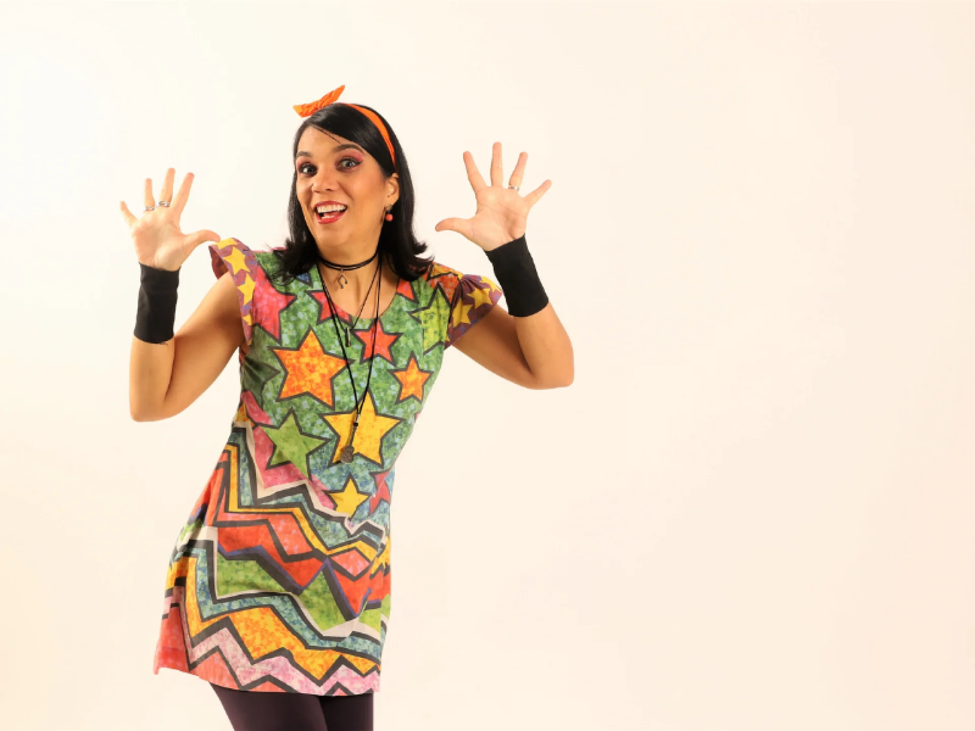 Mulher sorridente com vestido colorido de estampa geométrica e estrelas, levantando as duas mãos abertas. Ela usa uma tiara laranja e munhequeiras pretas, em fundo bege liso. Mulher sorridente com vestido colorido de estampa geométrica e estrelas, levantando as duas mãos abertas. Ela usa uma tiara laranja e munhequeiras pretas, em fundo bege liso.