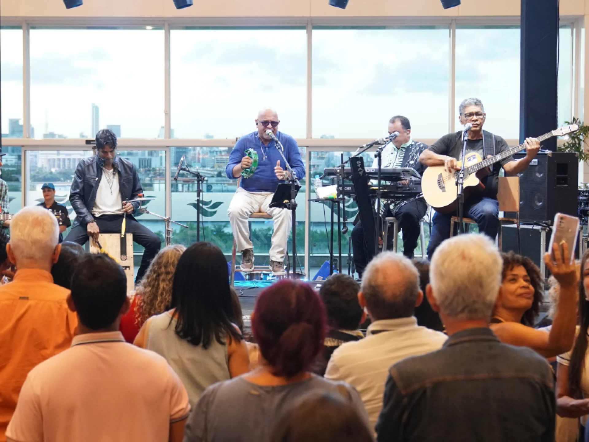 Uma banda se apresenta no palco com guitarra, teclado e microfone, enquanto uma multidão assiste do lado de dentro. Grandes janelas revelam uma paisagem urbana e um céu nublado ao fundo.
