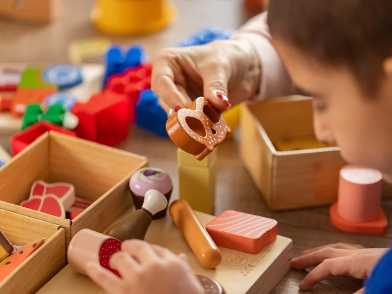 Criança e adulto exploram brinquedos de madeira que imitam alimentos, com blocos coloridos e caixinhas ao fundo.