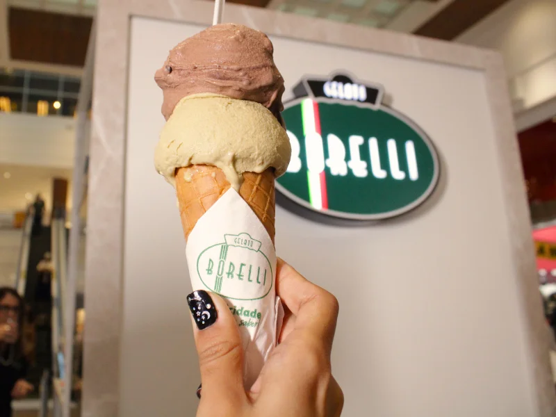 Uma mão com esmalte escuro segurando uma casquinha de sorvete com duas bolas de gelato (chocolate e pistache), com o logo da Borelli ao fundo.