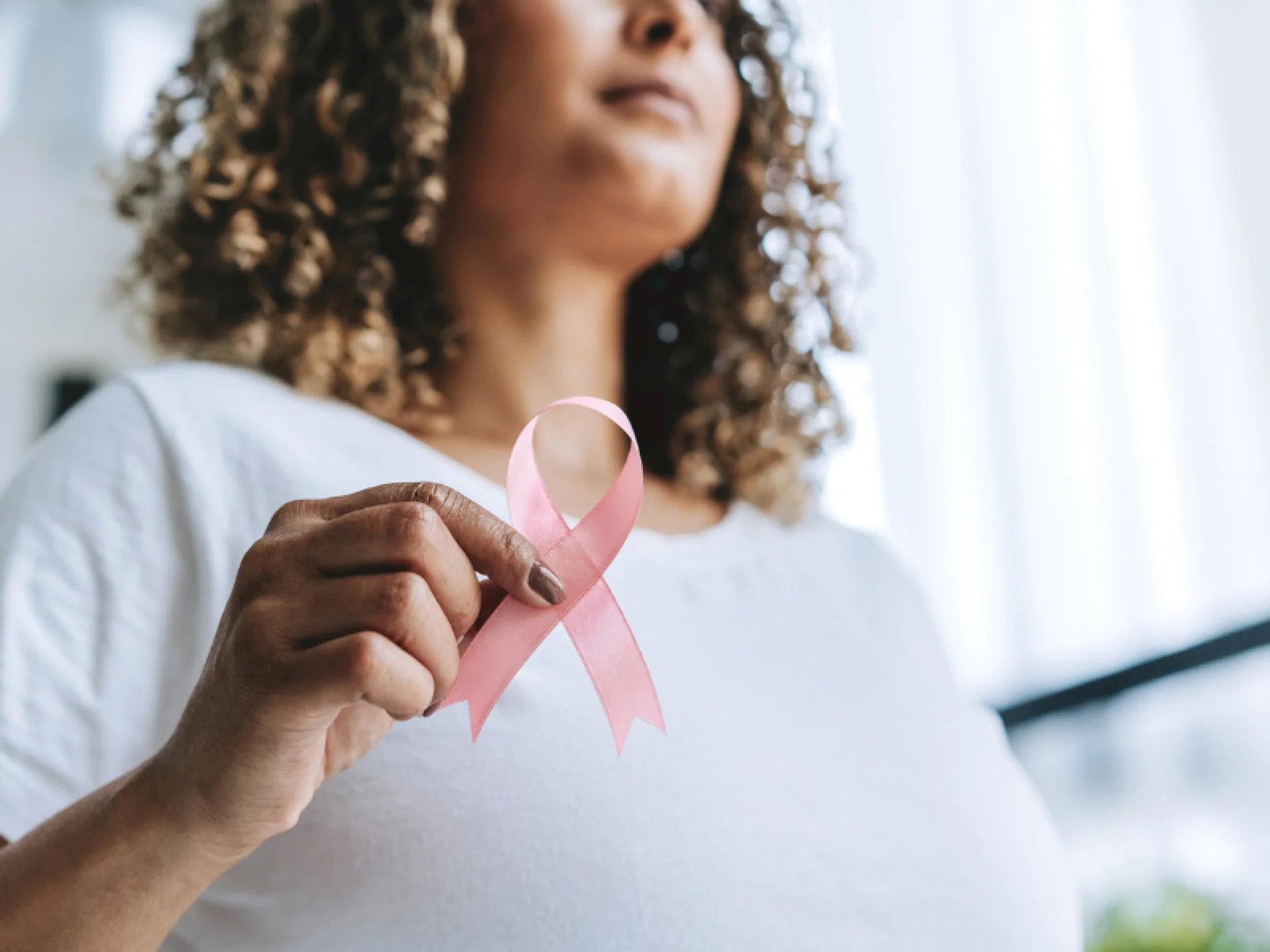 Mulher de cabelos cacheados, vestindo camiseta branca, segura um laço rosa (símbolo do combate ao câncer de mama) em frente ao peito. Mulher de cabelos cacheados, vestindo camiseta branca, segura um laço rosa (símbolo do combate ao câncer de mama) em frente ao peito.