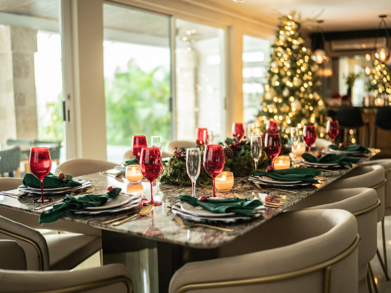 Mesa de jantar posta para o Natal, com taças vermelhas, guardanapos verdes, velas e arranjo de folhagens ao centro, cercada por cadeiras estofadas. Ao fundo, uma árvore de Natal iluminada em ambiente amplo com grandes portas de vidro.