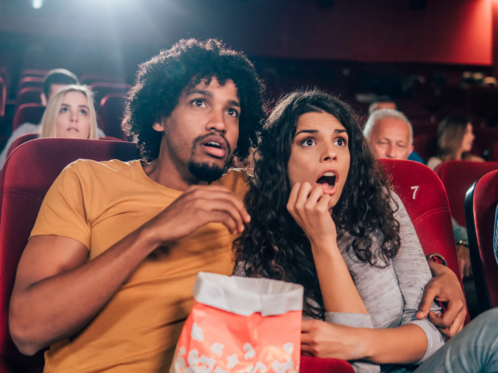 Casal sentados em poltronas do cinema comendo pipoca com expressão facial de surpresa.