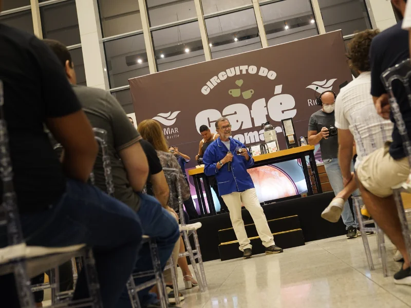 Romoaldo de Souza, um homem de cabelos e barba grisalhos vestindo dólmã azul e calça branca, fala ao microfone segurando uma xícara de café em um palco.