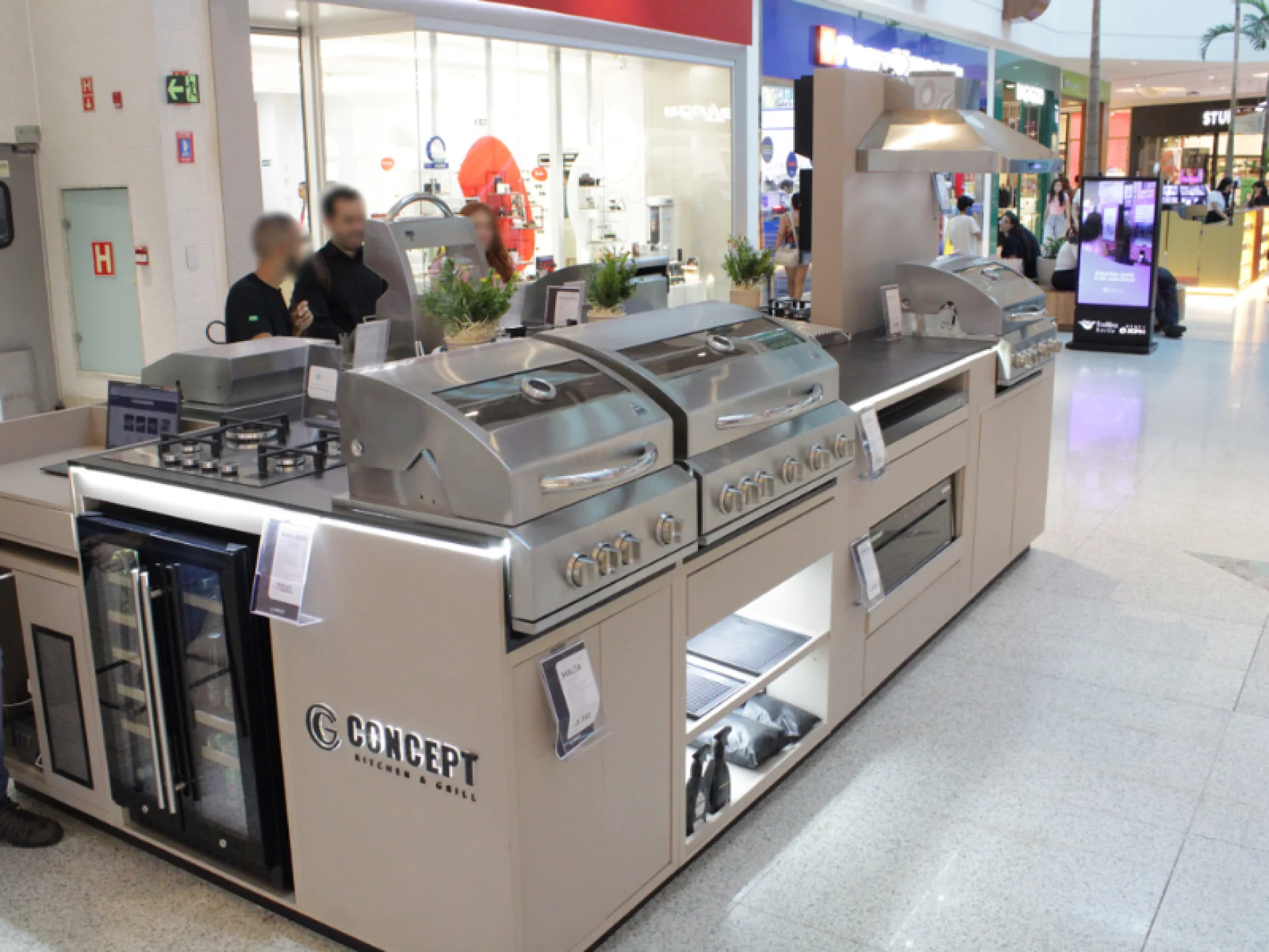 Visão geral de um estande de cozinha/churrasco modular em um shopping, com balcões beges e equipamentos em aço inoxidável. Inclui dois grandes grills com tampas, um fogão cooktop e um refrigerador de bebidas.