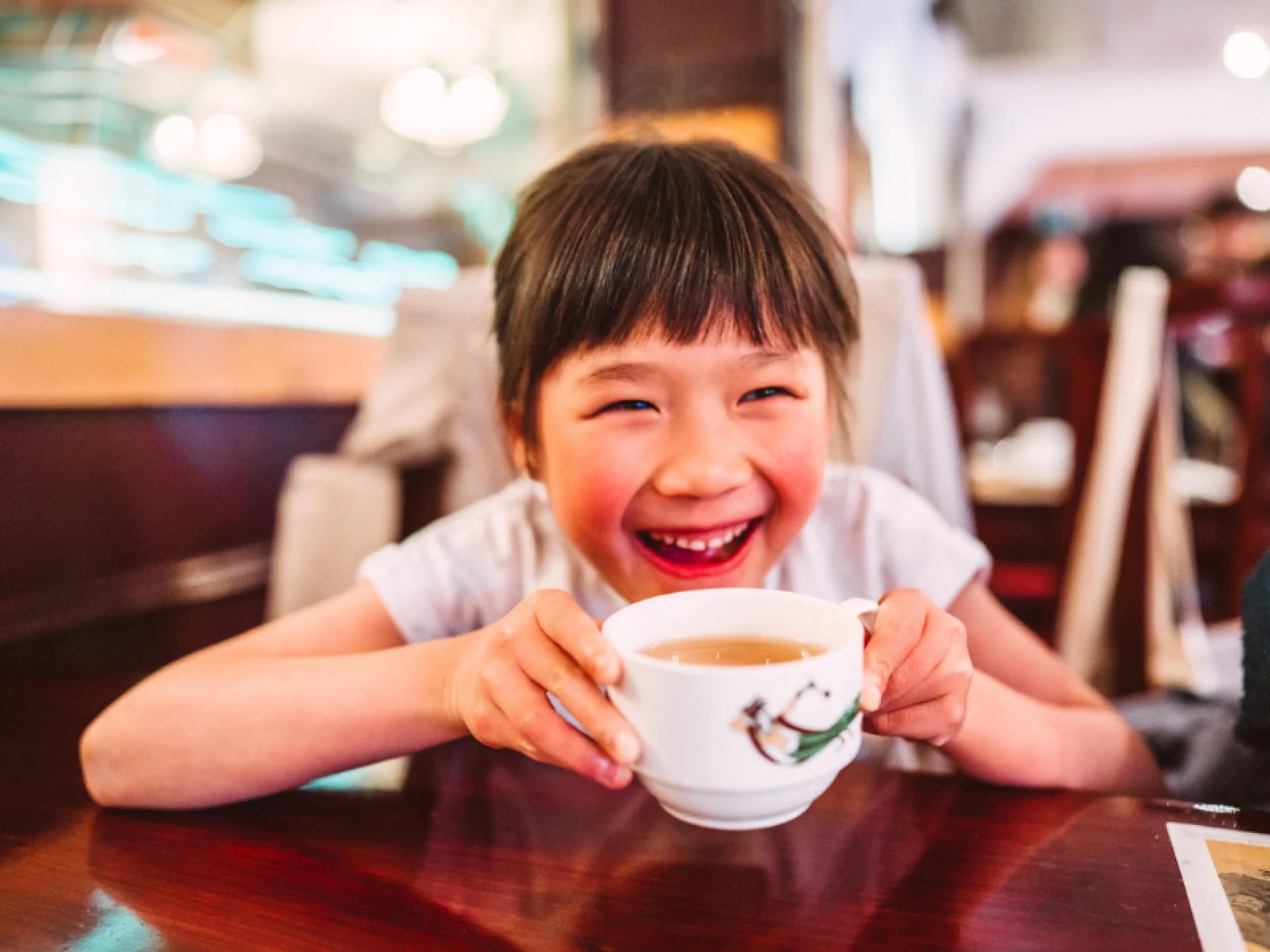 Criança segurando uma xícara contendo chá com os braços apoiados sobre a mesa. Criança segurando uma xícara contendo chá com os braços apoiados sobre a mesa.