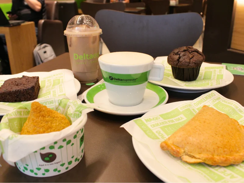 Uma variedade de salgados e doces da Deltaexpresso em uma mesa: um brownie, um muffin, uma coxinha no copo e um pastel assado, acompanhados de café quente e gelado.