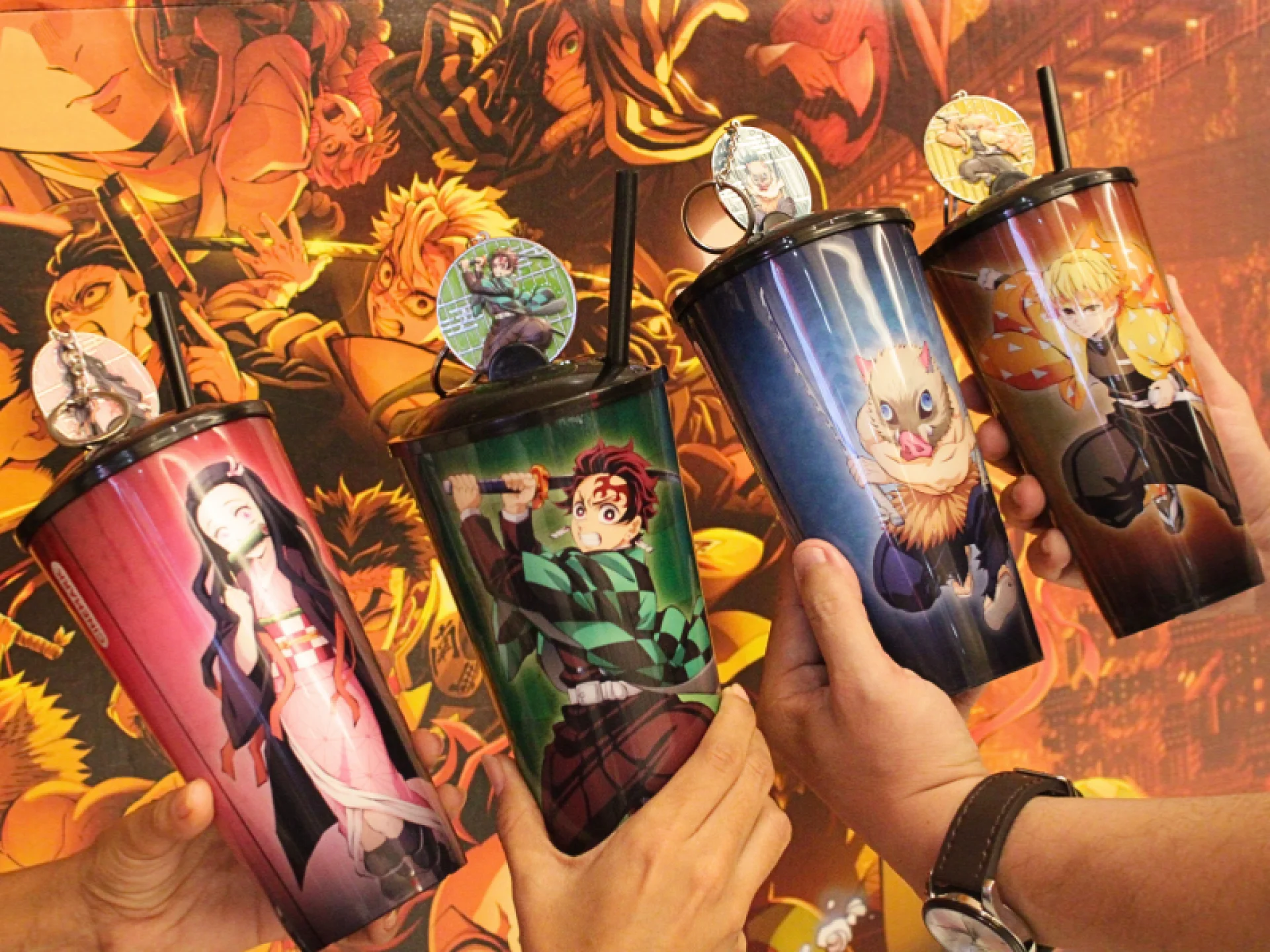 Quatro mãos seguram copos coloridos com desenhos de personagens do anime Demon Slayer. Cada copo apresenta um personagem diferente. O fundo mostra um pôster vibrante do anime.