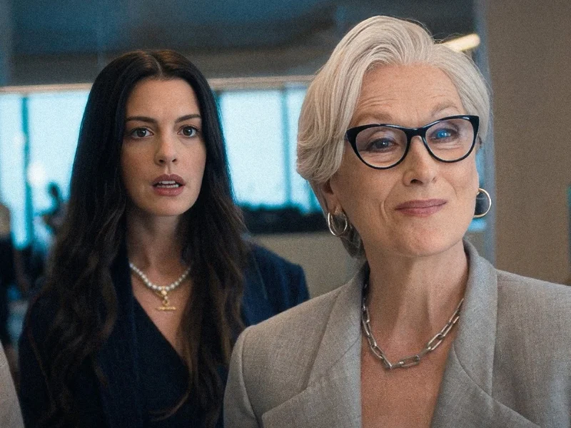 Cena do filme O Diabo Veste Prada 2 com Anne Hathaway e Meryl Streep em plano conjunto, Andrea elegante ao fundo e Miranda Priestly em destaque.
