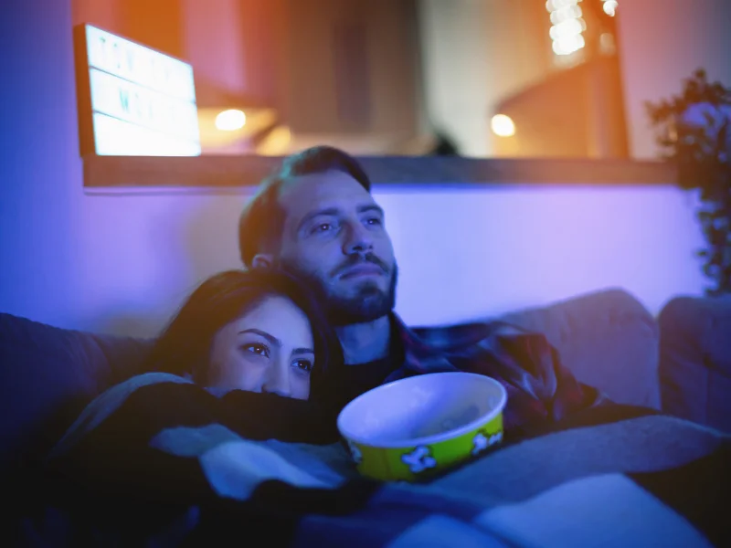 A imagem mostra um casal abraçado assistindo televisão com luzes escuras e um balde de pipoca