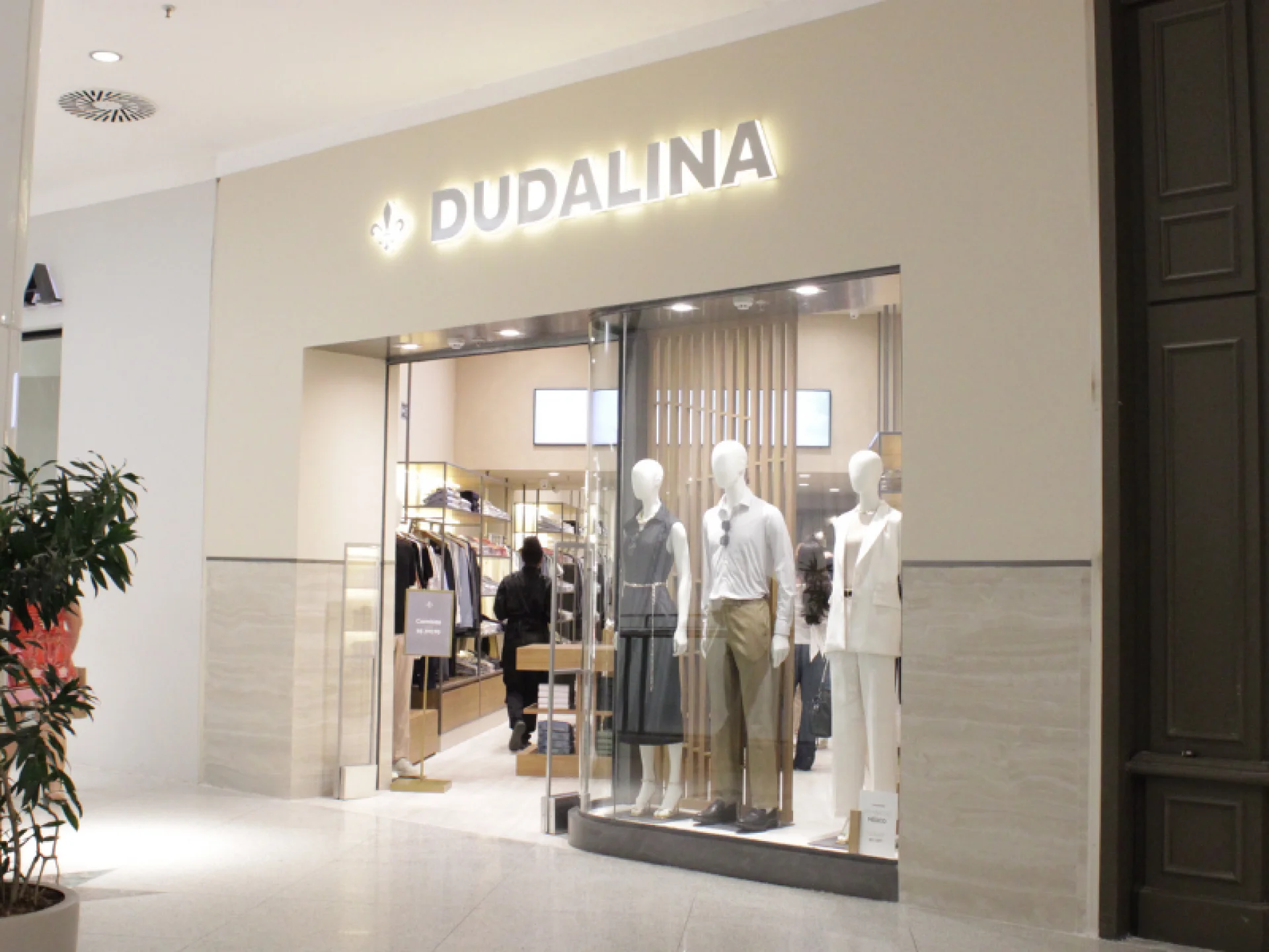 Fachada da loja Dudalina com o nome e o símbolo de flor-de-lis iluminados, exibindo manequins com roupas sociais na vitrine
