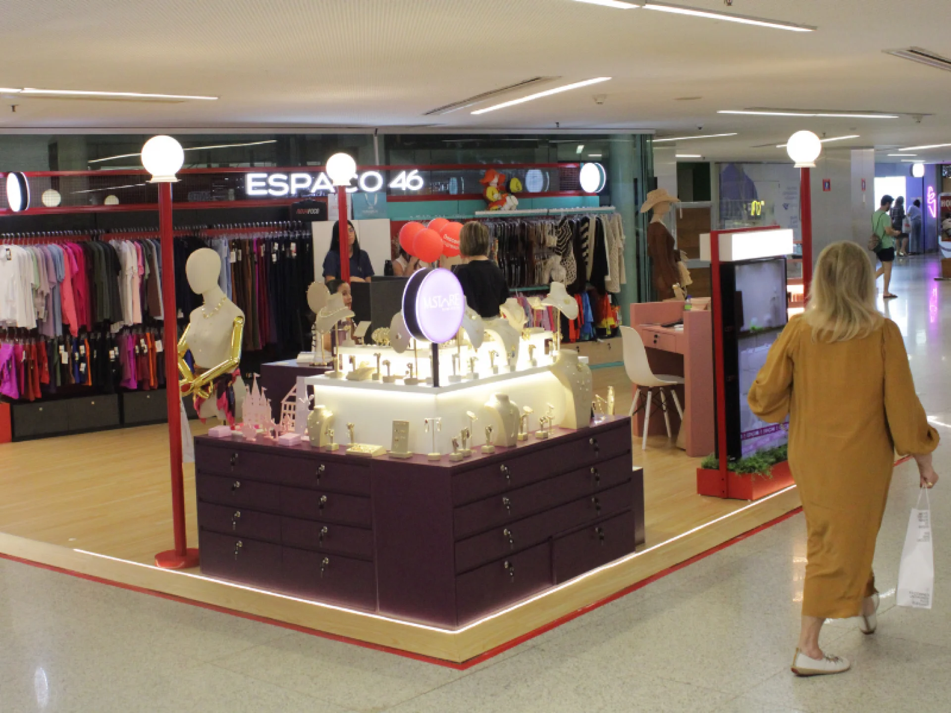 Vista angular de um quiosque de moda e acessórios iluminado. Há araras de roupas no fundo, um balcão central com joias e uma pessoa de vestido mostarda caminhando à direita.