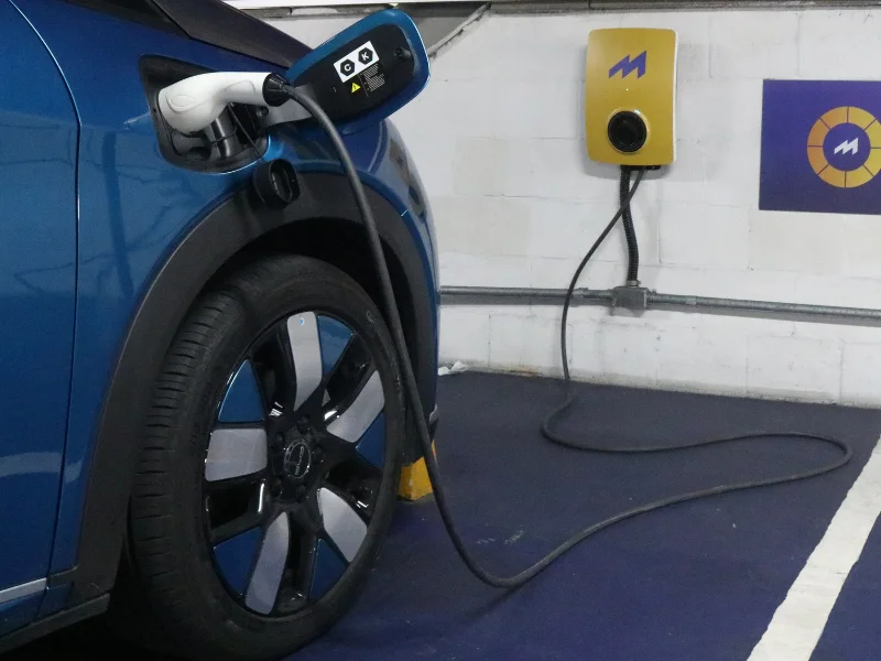 Um carro azul está estacionado em uma vaga de carregamento, com o cabo conectado à porta na lateral e a um carregador de parede amarelo-mostarda à direita. A foto mostra a roda, parte do carregador de bordo e o carregador fixo na parede de tijolos brancos.