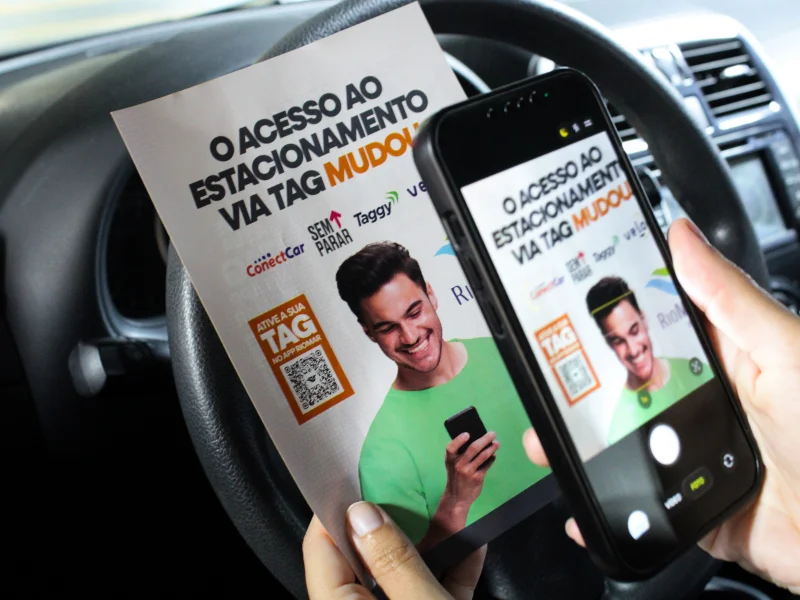 Mão segura folheto sobre nova tag de estacionamento à frente do volante, enquanto um celular aponta a câmera para o QR Code de ativação.