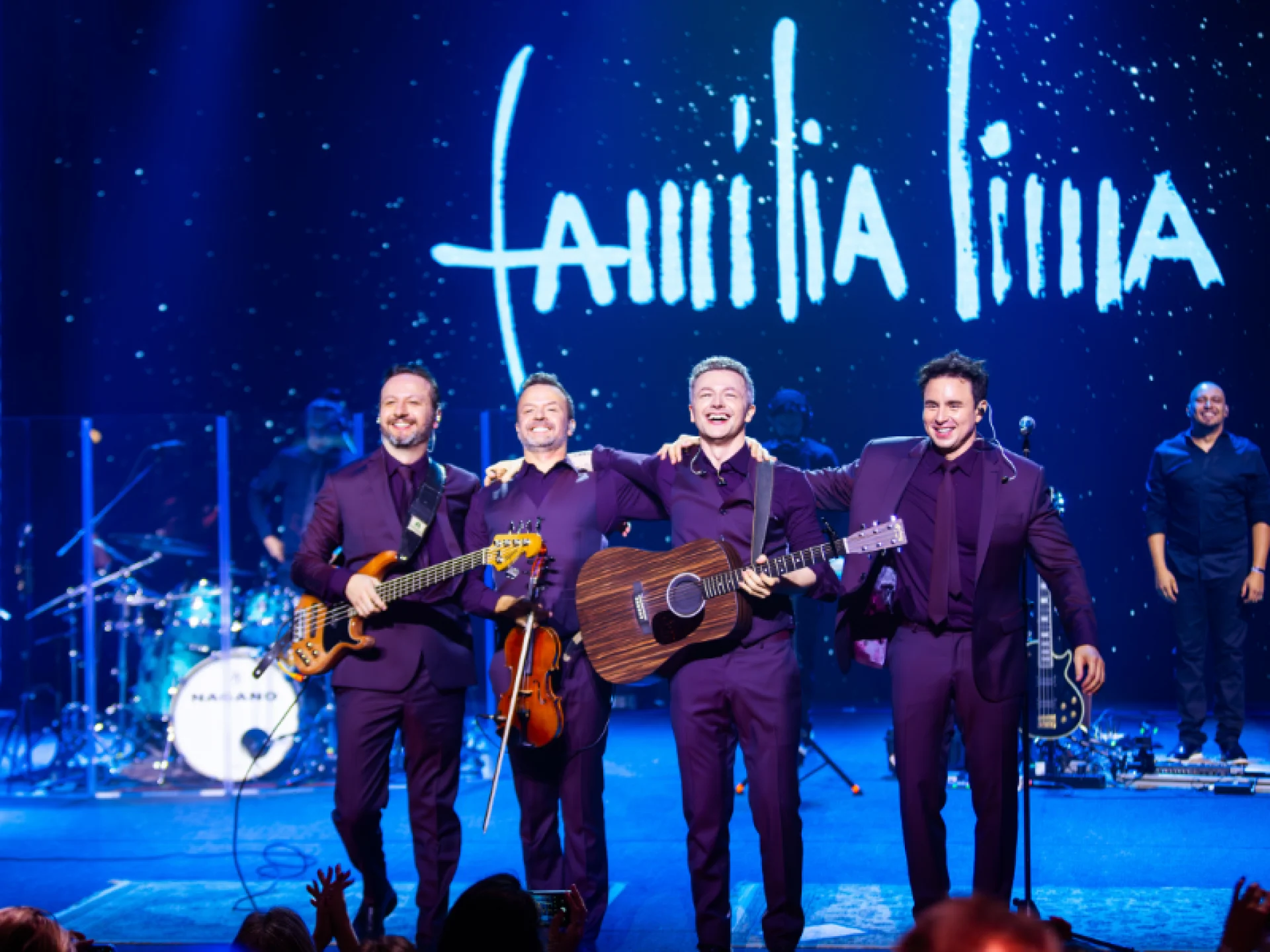 O grupo Família Lima, formado por quatro homens em ternos roxos, posa sorridente no palco segurando instrumentos musicais. Acima, o nome "Família Lima" é projetado em letras brancas. O grupo Família Lima, formado por quatro homens em ternos roxos, posa sorridente no palco segurando instrumentos musicais. Acima, o nome "Família Lima" é projetado em letras brancas.