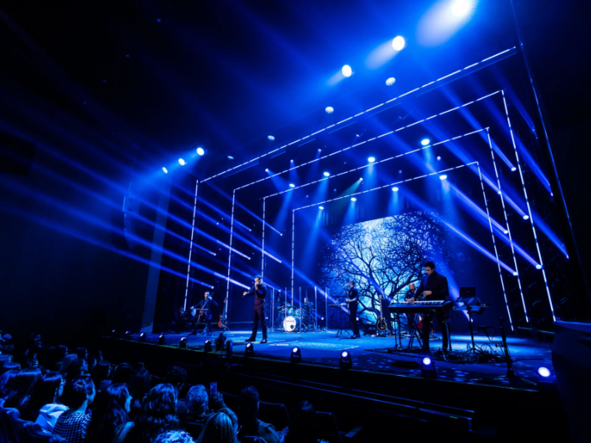 Palco de show com banda sob iluminação azul intensa, com focos de luz e linhas retangulares de LED. Ao centro, um vocalista canta em frente a um telão com a silhueta de uma árvore.