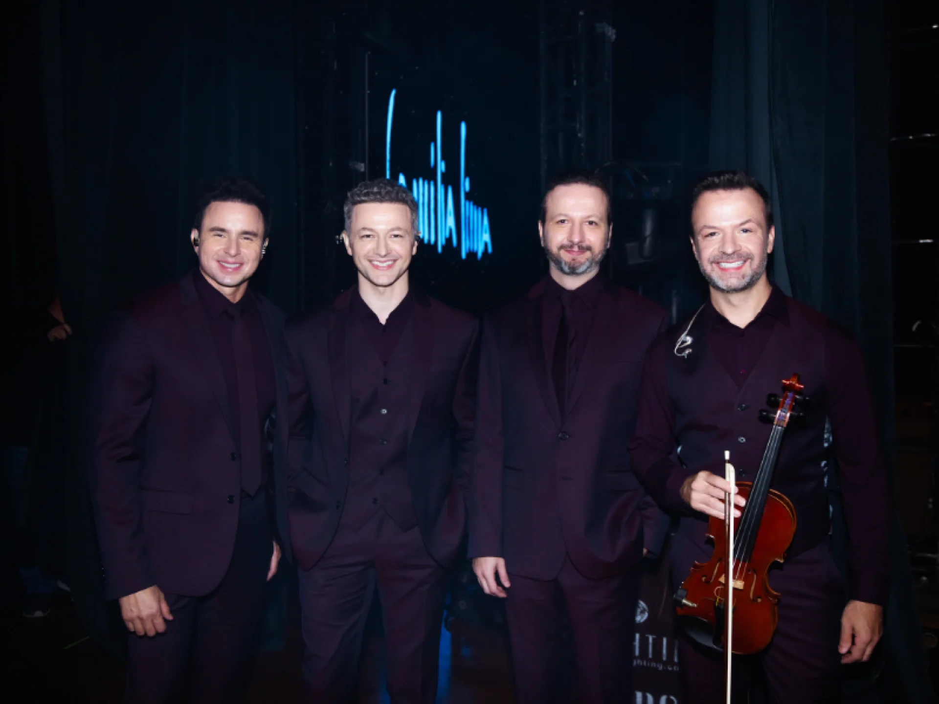 Os quatro integrantes da Família Lima posam lado a lado no palco, todos vestidos com ternos cor vinho escuro. Um deles segura um violino, e o fundo exibe o nome do grupo em luz neon azul. Os quatro integrantes da Família Lima posam lado a lado no palco, todos vestidos com ternos cor vinho escuro. Um deles segura um violino, e o fundo exibe o nome do grupo em luz neon azul.