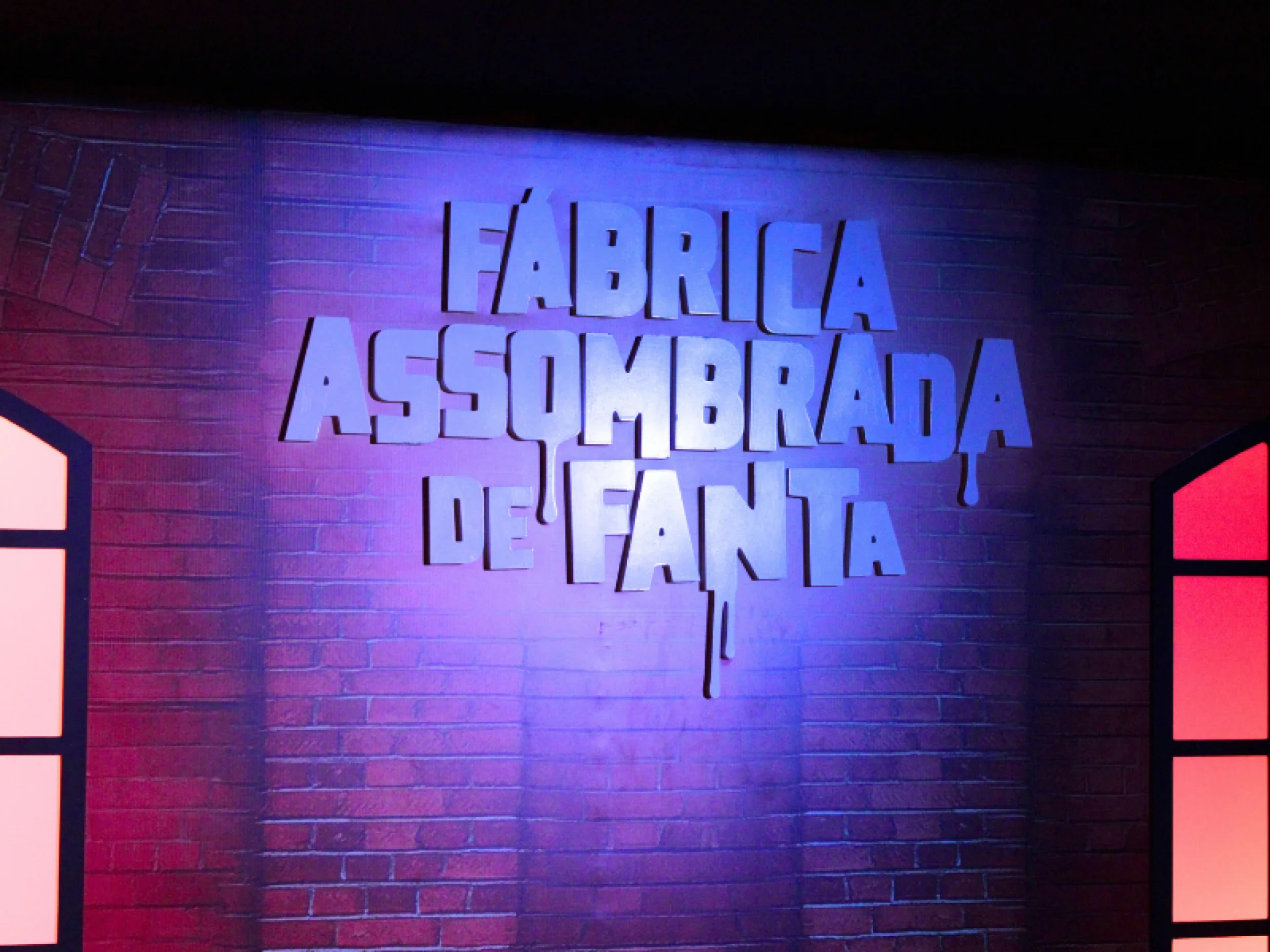 Um letreiro em relevo com "FÁBRICA ASSOMBRADA DE FANTA" é iluminado por uma luz roxa em uma parede de tijolos escuros. O design das letras inclui um efeito de líquido escorrendo.