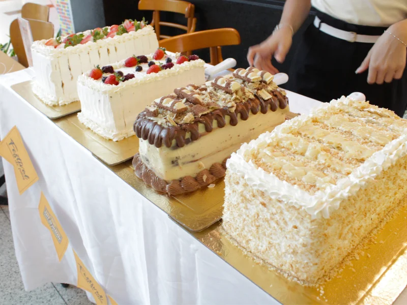 Mesa com quatro bolos retangulares expostos: dois brancos com frutas vermelhas frescas, um com cobertura de creme e pedaços de chocolate, e um coberto com coco ralado e chantilly.