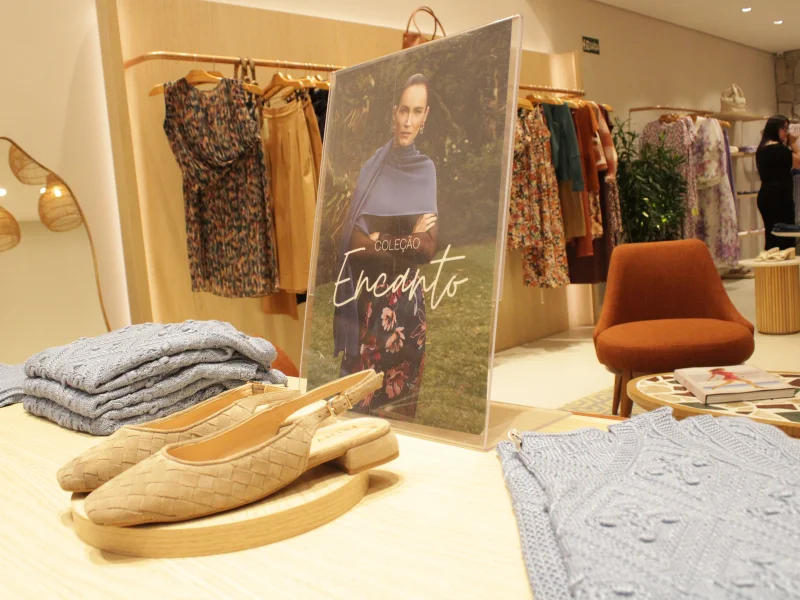 Display de loja com sapatos de bico fino trançados e placa da "Coleção Encanto" em um ambiente clean e moderno.