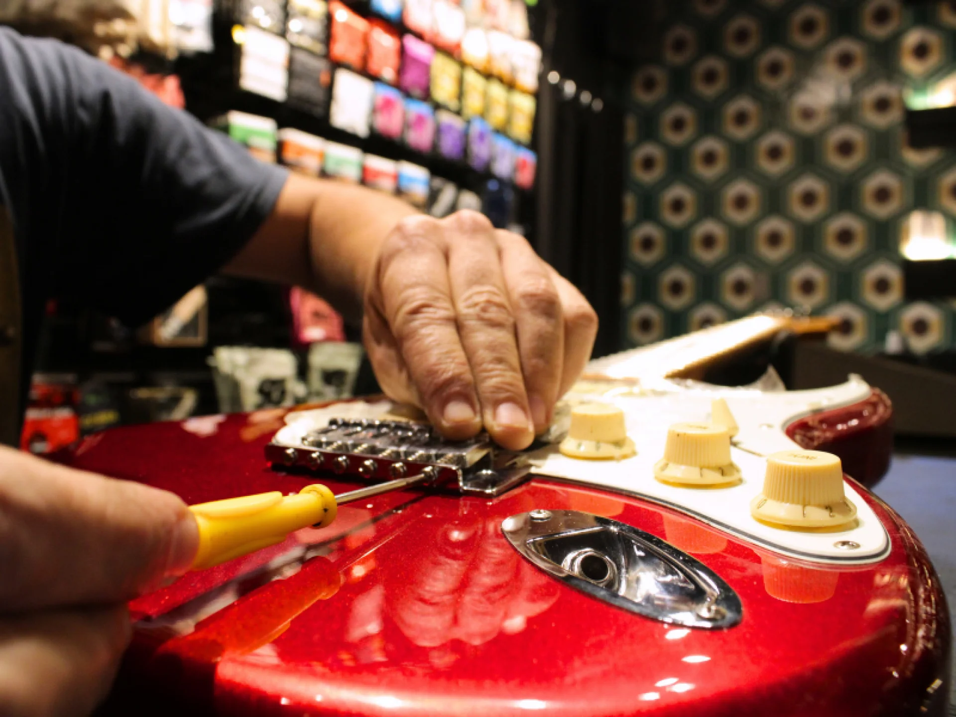 Mãos sobre a guitarra com uma ferramenta realizando conserto no instrumento com um fundo desfocado.