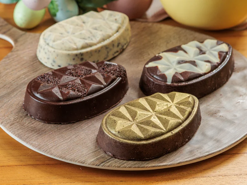 Visão geral de quatro ovos de chocolate artesanais com diferentes texturas e acabamentos sobre uma tábua de madeira.