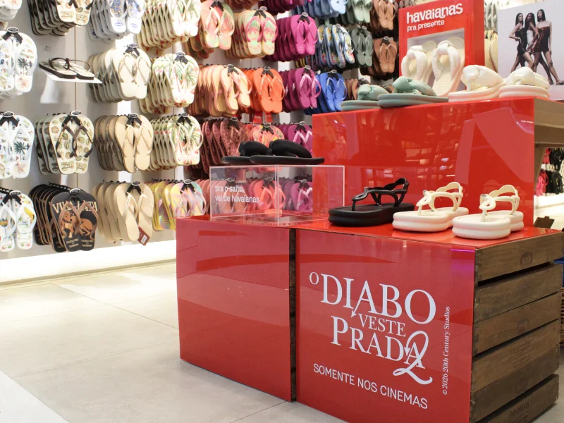 Expositor de loja com o logotipo do filme "O Diabo Veste Prada", apresentando diversos modelos de sandálias e chinelos modernos da coleção.