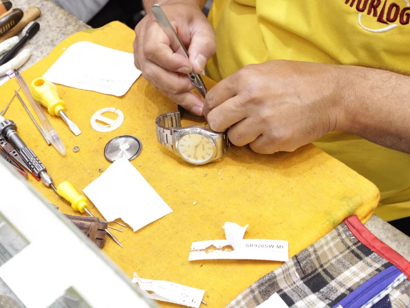 A imagem mostra as mãos de um homem consertando um relógio, numa bancada amarela. Ele usa a farda da horloge na cor amarela.