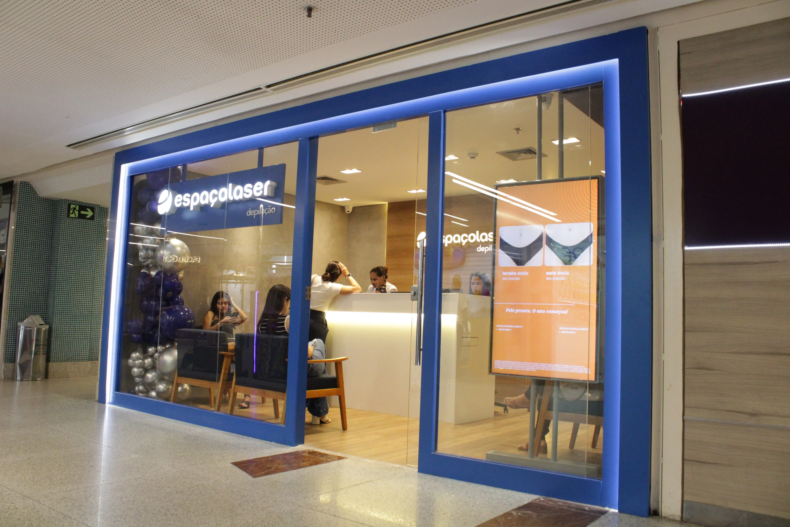 Fachada da Espaçolaser, com moldura azul iluminada, interior clean e clientes aguardando no balcão e poltronas. Um painel digital à direita exibe resultados de depilação, enquanto balões decorativos ocupam o canto esquerdo da vitrine de vidro.