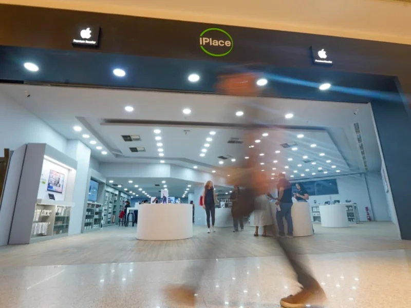 Fachada de uma loja iPlace no RioMar Recife, com iluminação moderna e ampla abertura frontal. Clientes circulam pelo espaço enquanto um pedestre passa em movimento na frente da loja.