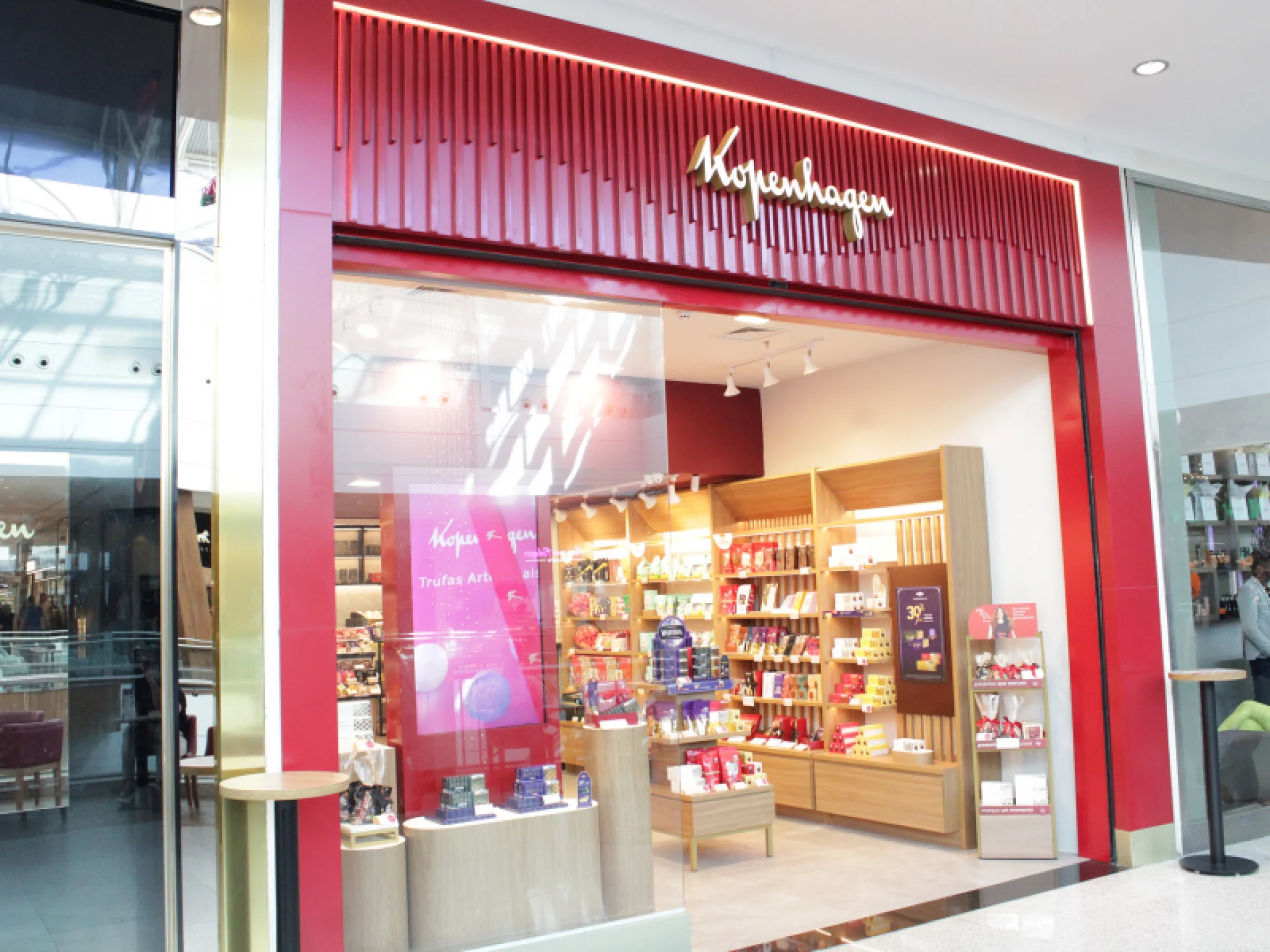 Fachada de uma loja Kopenhagen em um shopping, com uma entrada de vidro emoldurada em vermelho escuro. O interior da loja exibe prateleiras de madeira clara e produtos de chocolate.