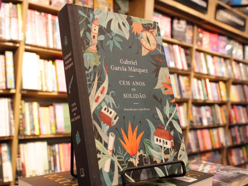 A foto exibe uma edição especial ilustrada do clássico "Cem Anos de Solidão". A capa é de um tom verde-azulado escuro e tem uma arte detalhada no estilo botânico/tropical, com elementos do realismo mágico.