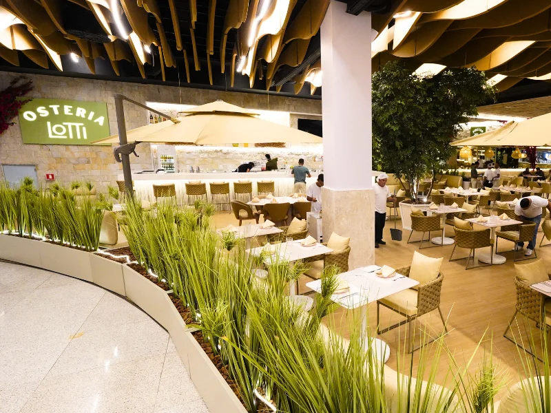 Visão ampla do restaurante com teto moderno ondulado, bar iluminado ao fundo e mesas distribuídas entre plantas e ombrelones.