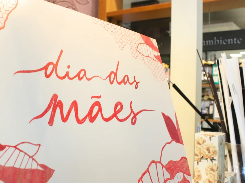 Placa branca com a frase "dia das mães" escrita em caligrafia vermelha, com detalhes florais nas bordas, em primeiro plano na vitrine da loja.