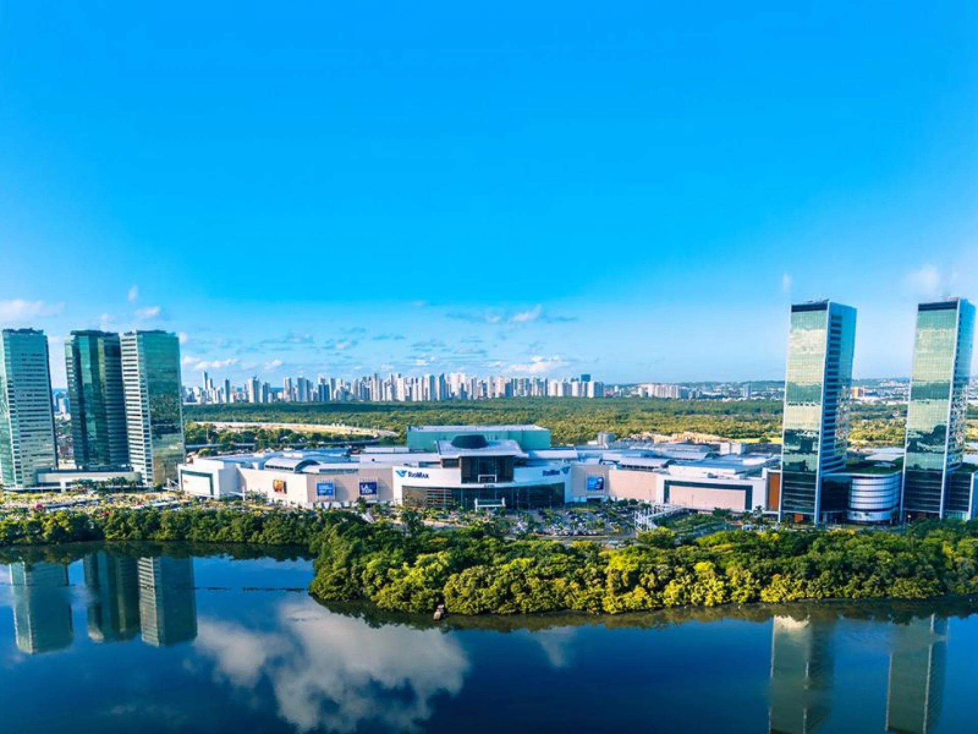 A imagem mostra uma foto do RioMar Recife a longa distância em um dia ensolarado com o céu azul. A foto também mostra as três torres empresariais do lado esquerdo e as outras duas torres do lado direito. A imagem mostra uma foto do RioMar Recife a longa distância em um dia ensolarado com o céu azul. A foto também mostra as três torres empresariais do lado esquerdo e as outras duas torres do lado direito.