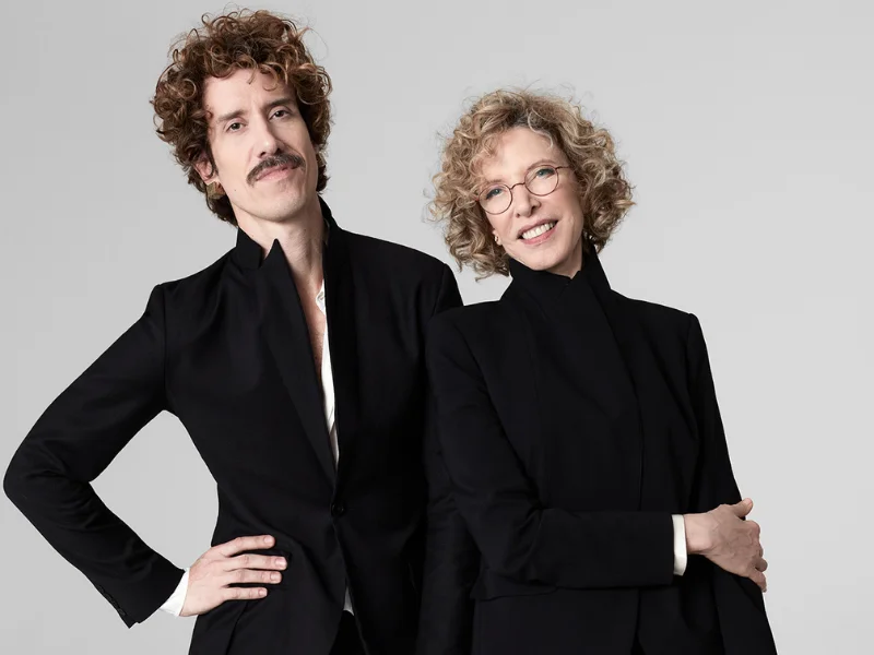 Marília Gabriela e seu filho, Theodoro Cochrane, posam lado a lado vestindo blazers pretos elegantes, ambos exibindo cabelos cacheados e um visual sofisticado sobre um fundo neutro.