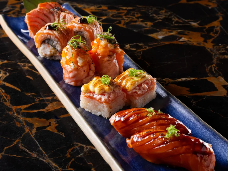 Travessa longa com variados sushis de salmão levemente selados no fogo, finalizados com cremes, molhos escuros e cebolinha.