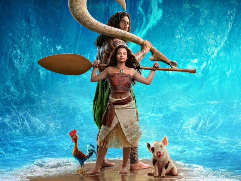 Live-action de Moana e Maui em pé na praia, ao lado do oceano azul-turquesa; ela segura um remo de madeira, um galo e um leitão aparecem aos pés dos dois.