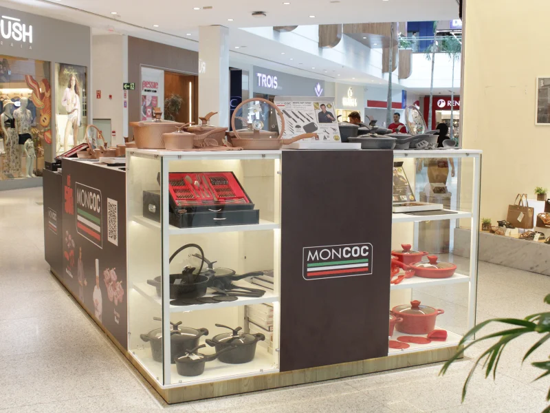 Quiosque de shopping da marca "Moncoc" exibindo conjuntos de panelas em cores variadas (cobre, grafite e vermelho) em prateleiras de vidro iluminadas e no balcão superior.