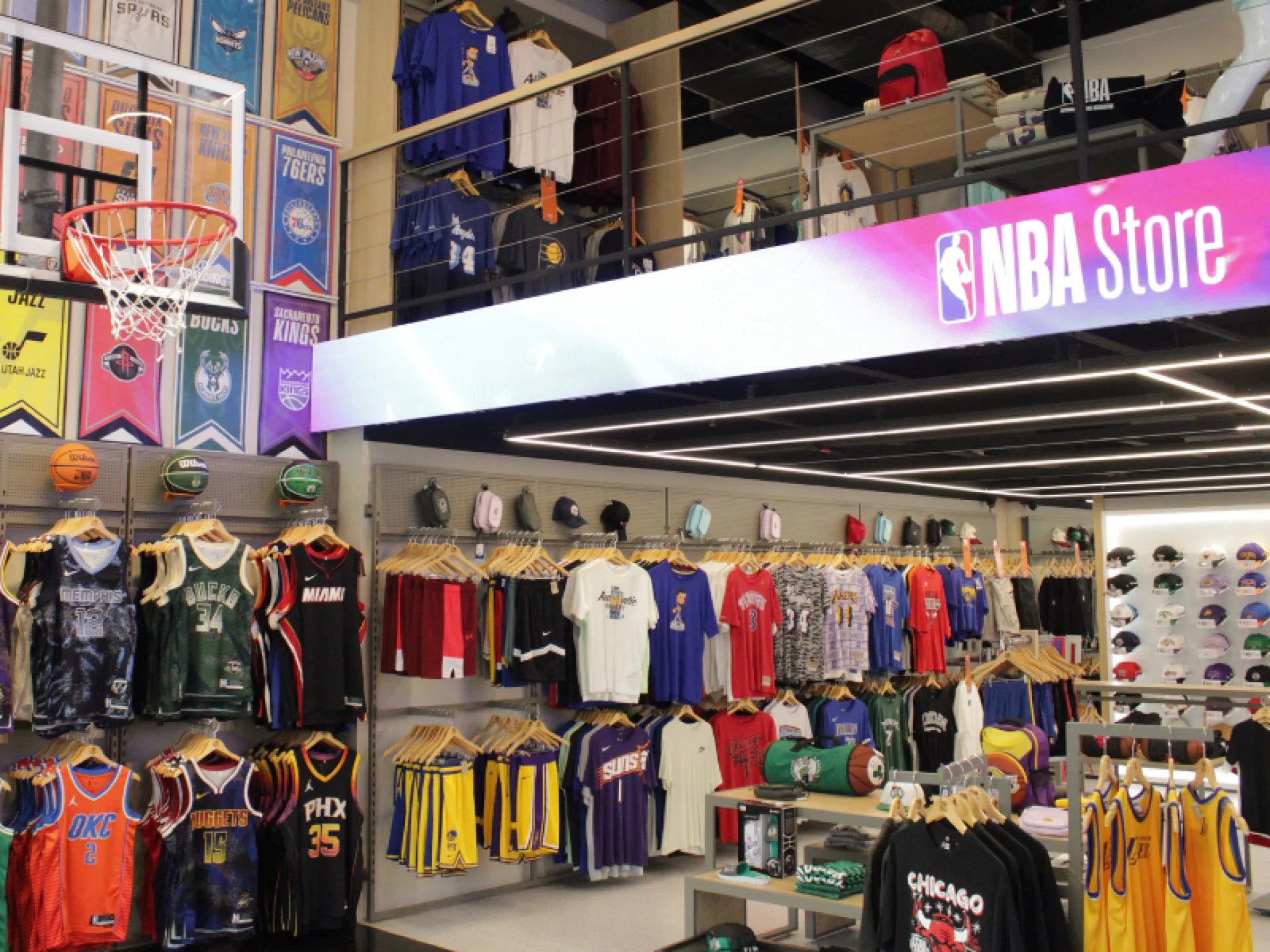 A imagem mostra o interior de uma loja da NBA, com camisas penduradas de times variados, uma cesta de basquete e banners de times no andar superior. A imagem mostra o interior de uma loja da NBA, com camisas penduradas de times variados, uma cesta de basquete e banners de times no andar superior.