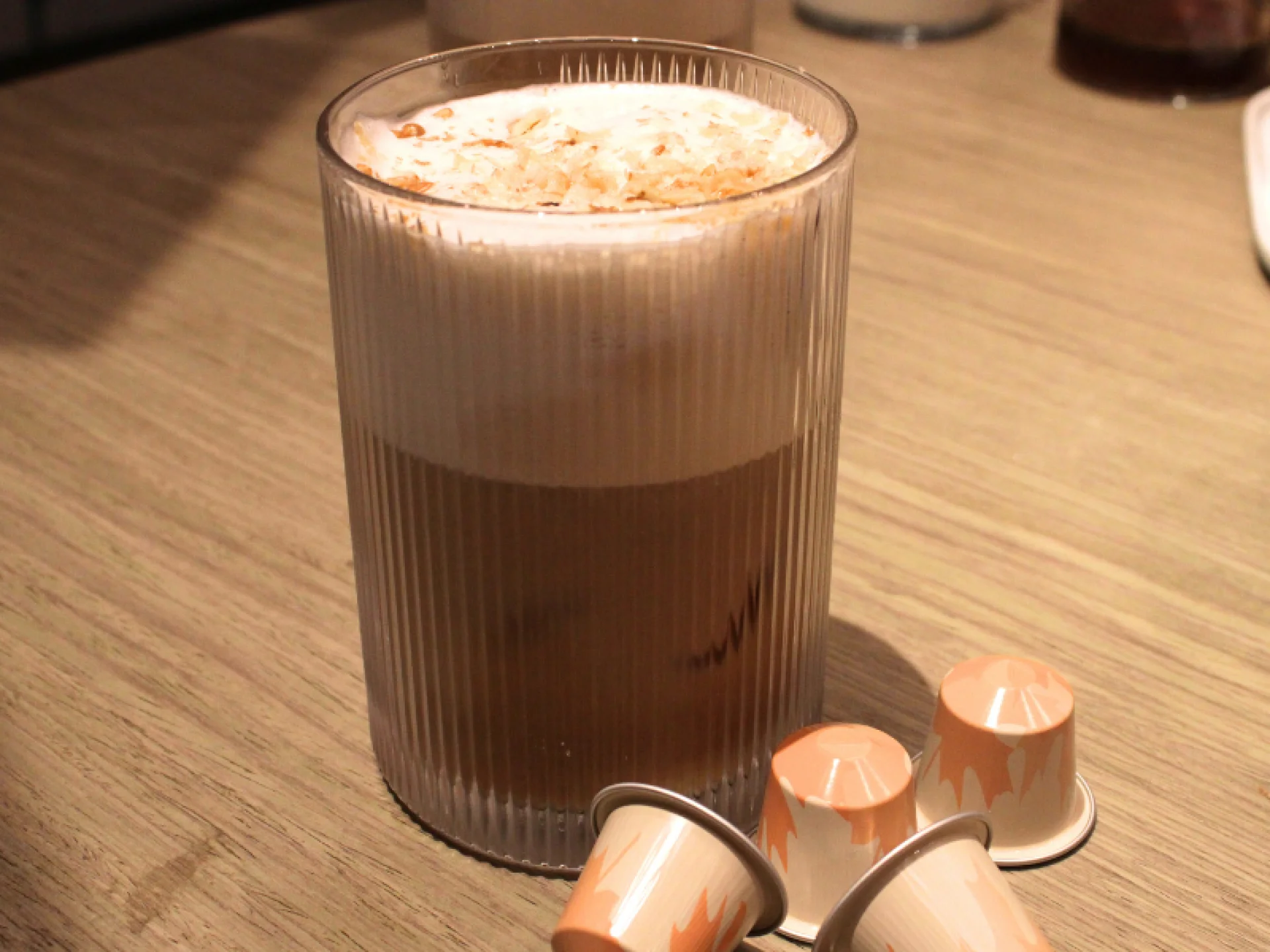 A imagem mostra um café gelado finalizado com espuma e nozes raladas, com cápsulas Nespresso laranjas espalhadas na base. A imagem mostra um café gelado finalizado com espuma e nozes raladas, com cápsulas Nespresso laranjas espalhadas na base.
