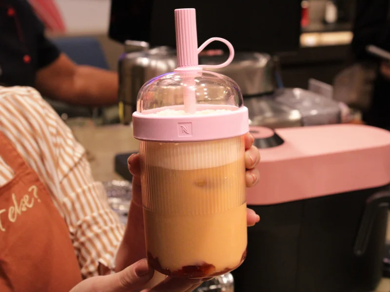Pessoa segura um copo portátil Nespresso rosa e transparente com uma bebida gelada de café em camadas e um canudo estruturado.