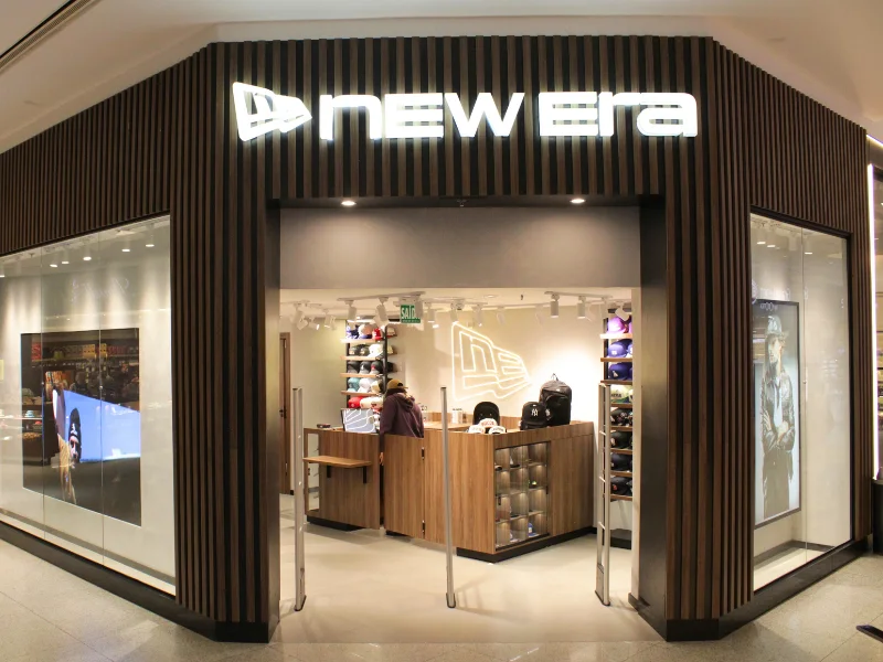 Fachada frontal da loja New Era em um shopping, apresentando letreiro luminoso e interior moderno com acabamento em madeira.