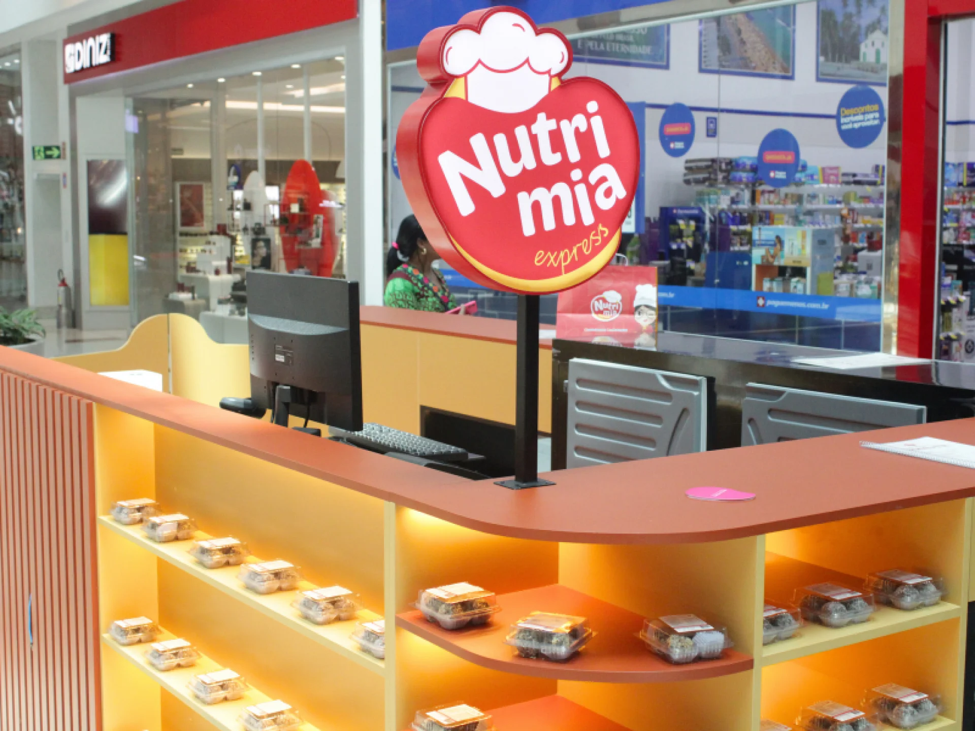 A imagem mostra o quiosque da Nutrimia na cor laranja com luzes amarelas e com a logo nas cores vermelho e branco A imagem mostra o quiosque da Nutrimia na cor laranja com luzes amarelas e com a logo nas cores vermelho e branco