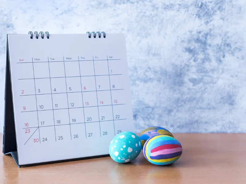 Calendário de mesa branco marcando o mês de abril ao lado de três ovos de Páscoa coloridos e pintados à mão sobre uma mesa de madeira.
