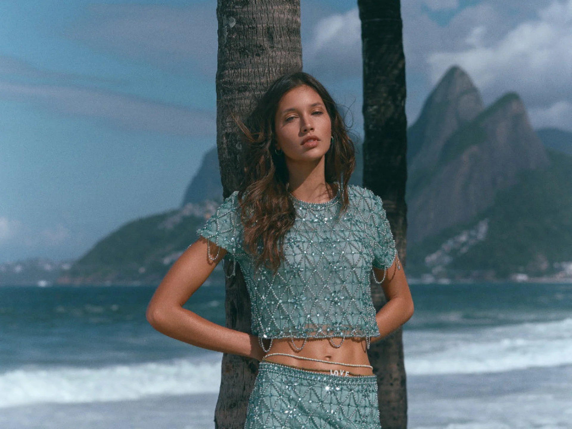 Uma modelo com top cropped e minissaia turquesa cravejados de mi&ccedil;angas posa na praia, com coqueiros e o Morro Dois Irm&atilde;os ao fundo.