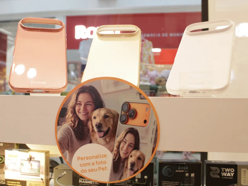 Três capas de celular em tons pastéis (rosê, bege e branco) estão expostas sobre um adesivo redondo que mostra uma mulher com seu cachorro, promovendo a personalização com fotos de pets.